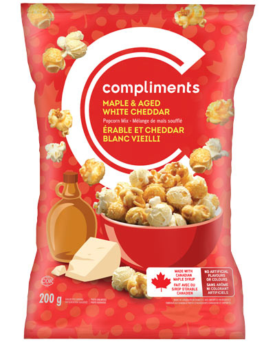 Red package of Compliments Maple & Aged White Cheddar Popcorn Mix sitting in a red bowl on package alongside a wedge of cheddar and a bottle of maple syrup.