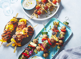 Three kinds of European-inspired kabobs on three different platters: one that’s gray, blue and white with water glasses in the background and a blue napkin.