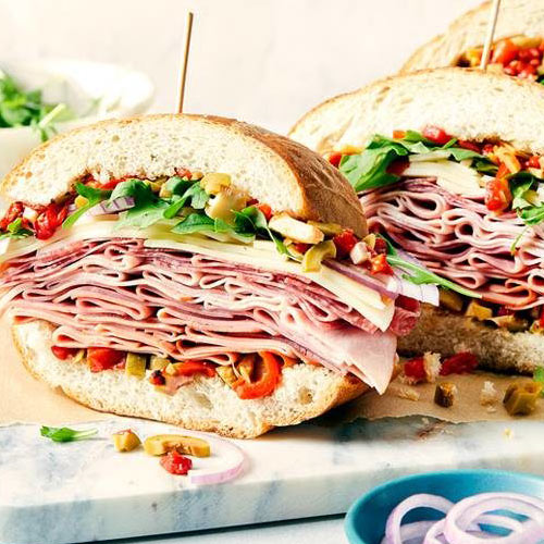 A sky high deli meat, cheese, olive, roasted peppers, red onion and arugula filled French loaf muffuletta sitting on a marble chopping block with next to a bowl of chopped, pickled vegetables.