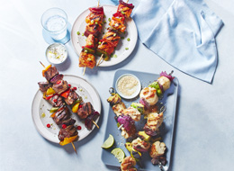 Three North-American-flavoured skewers including one beef, one chicken and one salmon on blue and white and gray plates with water glasses off to the side.