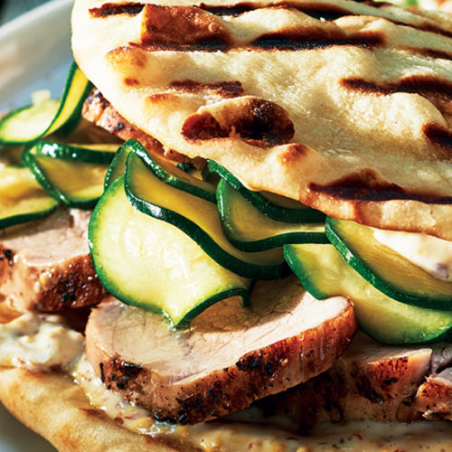 Grilled naan bread sandwiching grilled pork medallions and quick zucchini pickles sitting on a white plate next to a side of coleslaw.