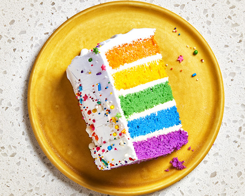 Sliced side shot of a piece of bakery rainbow cake with its multicoloured layers, iced in white and sitting atop a bright-yellow plate.