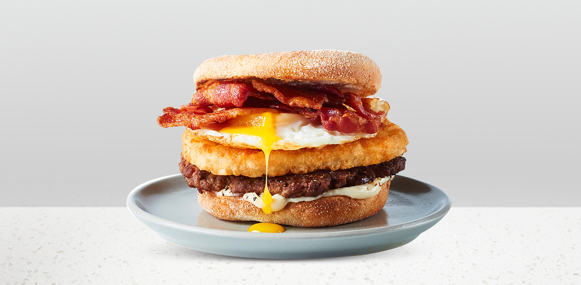 Smash burger on an English muffin topped with a fried egg, hash browns, bacon and a drizzle of maple syrup on a blue gray plate.