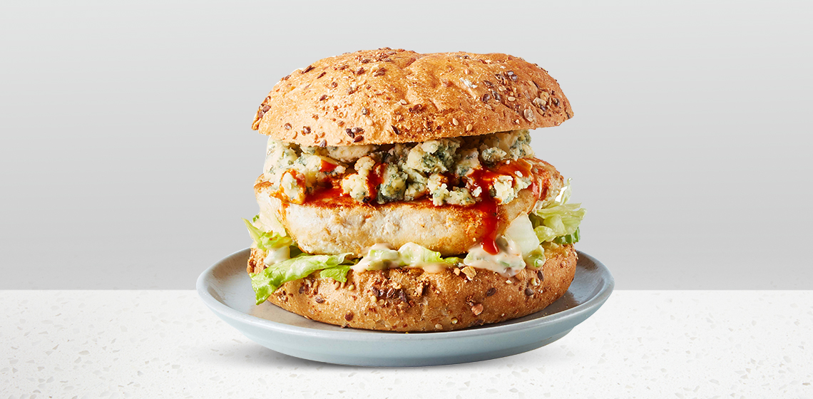 Lean chicken burger patty sandwiched in a seeded bun, topped with hot sauce, iceberg lettuce, Buffalo mayonnaise and blue cheese sitting on a blue gray plate.