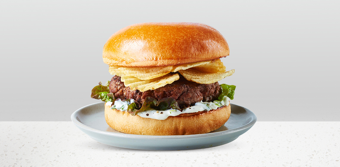 Fresh angus burger topped with ripple potato chips, red leaf lettuce and onion dip spread sitting on a blue gray plate. 