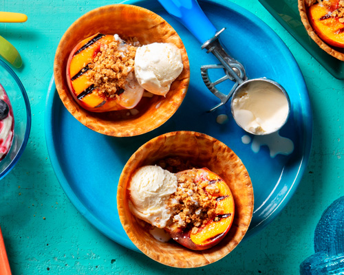 Two grilled peach crisps topped with oat crumble sitting on a blue plate and one sitting on a dark green plate with an ice cream scoop.