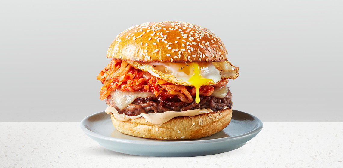 Sesame bun topped with a Compliments pork burger patty, fried egg, kimchi, kimchi mayonnaise and cheese sitting on a blue gray plate.