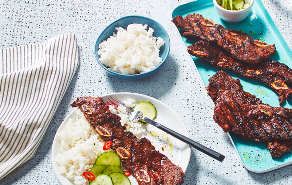 Short ribs on a blue plate with a complete, served plate next to it with rice and pickles.