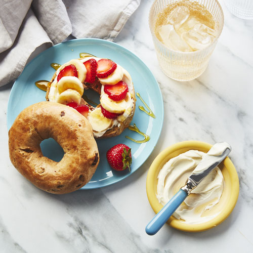 Cinnamon swirled bagel topped with Greek yogurt, cream cheese, strawberry and banana slices and a drizzle of honey on a pale blue plate.