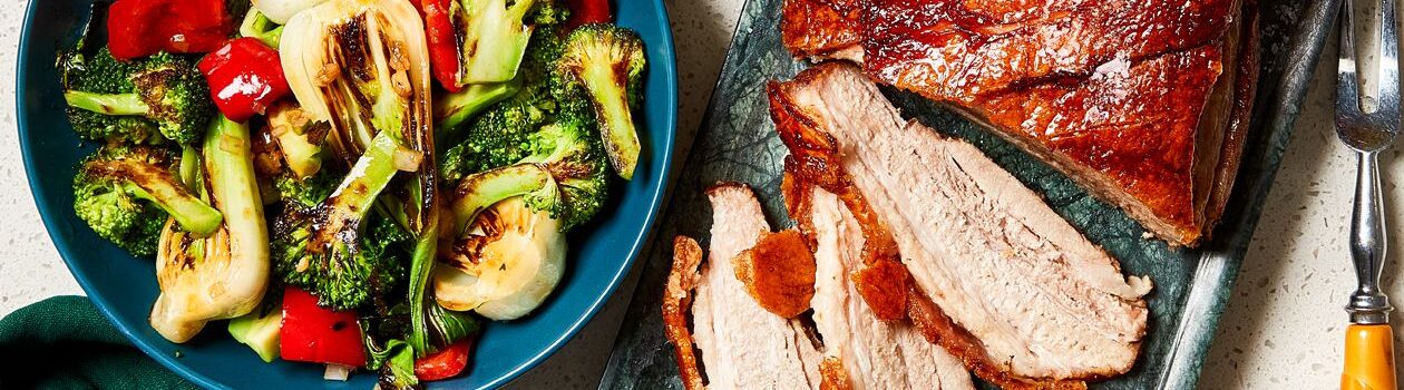 Slices of crispy pork belly on a gray platter with a bowl of charred bok choy and broccoli beside it in a blue bowl on a white countertop.