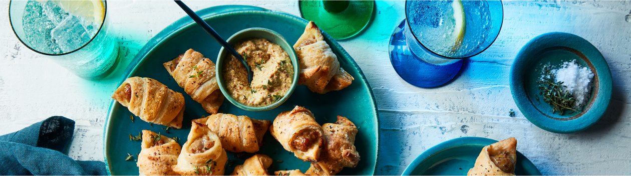 Tablescape with sparkling waters, napkin and round aqua green-blue serving plate with bratwurst pigs in blankets and a small bowl of grainy Dijon mustard for serving