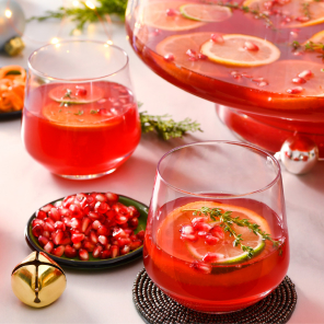 Four glasses of Rudolph Punch, a large punch bowl of Rudolph Punch and a dish of pomegranate seeds on a holiday-themed table