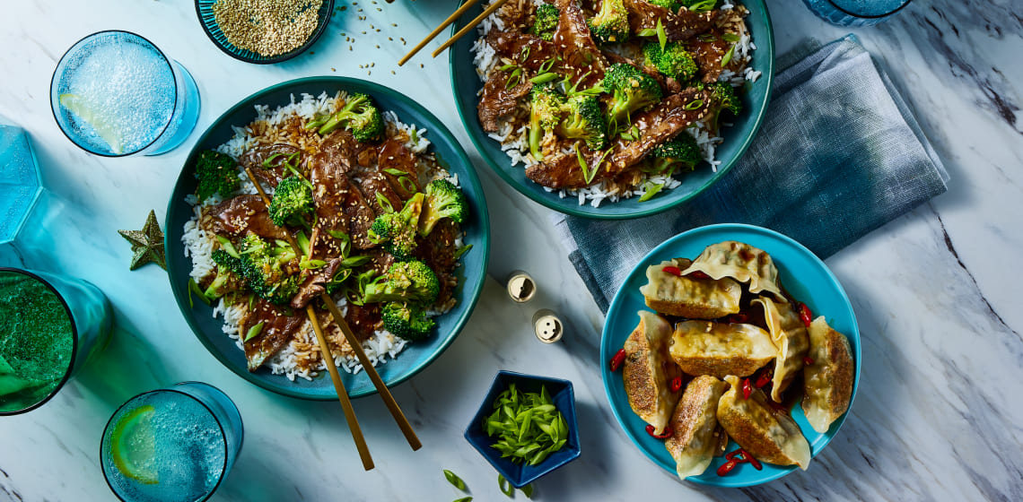 Two plates of beef and broccoli on a bed of rice with a side of potstickers