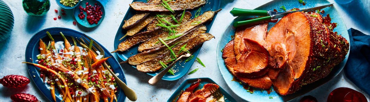 Light blue table surface with three serving platters featuring glazed ham, roasted eggplant and middle-eastern-inspired carrots with pomegranate and yogurt sauce.