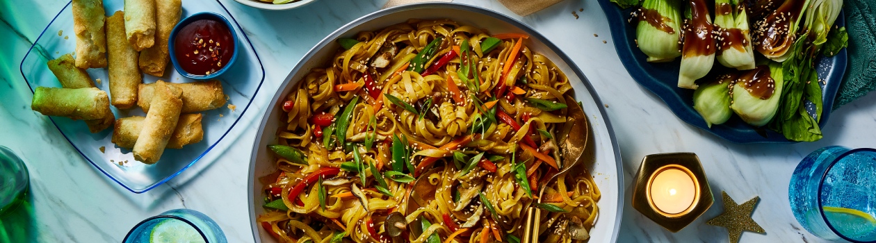 Bowl of vegetable Singapore noodles with a side of spring rolls and bok choy.