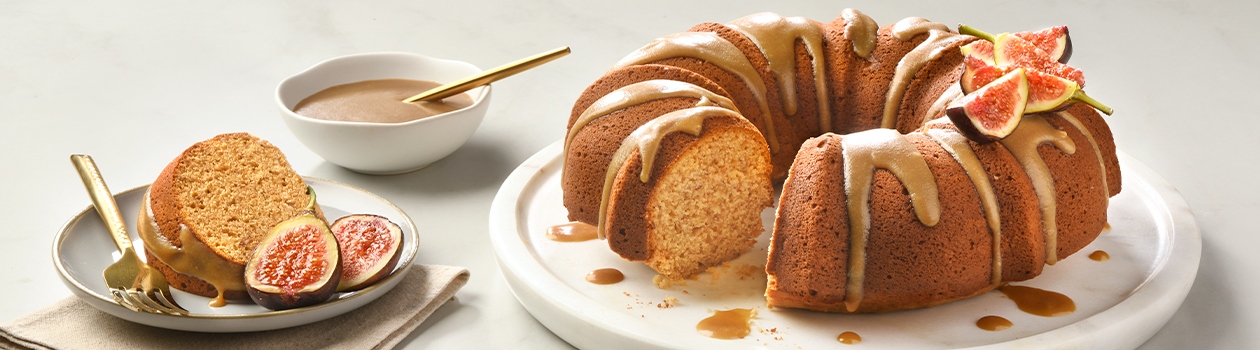 A bundt cake with a stick toffee glaze and fresh figs on top, a bowl of the toffee glaze, and a slice on another plate with a fork and a couple pieces of fig.