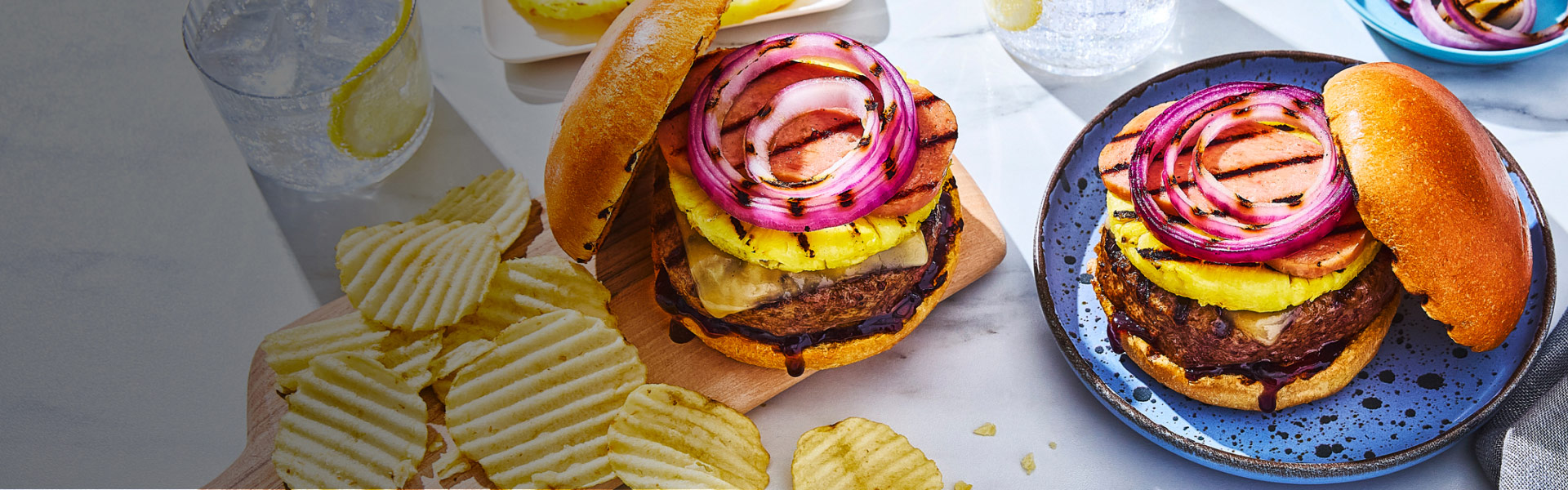 Two Hawaiian-inspired burgers on a white background with grilled pineapples and chips and two glasses of lemon water.