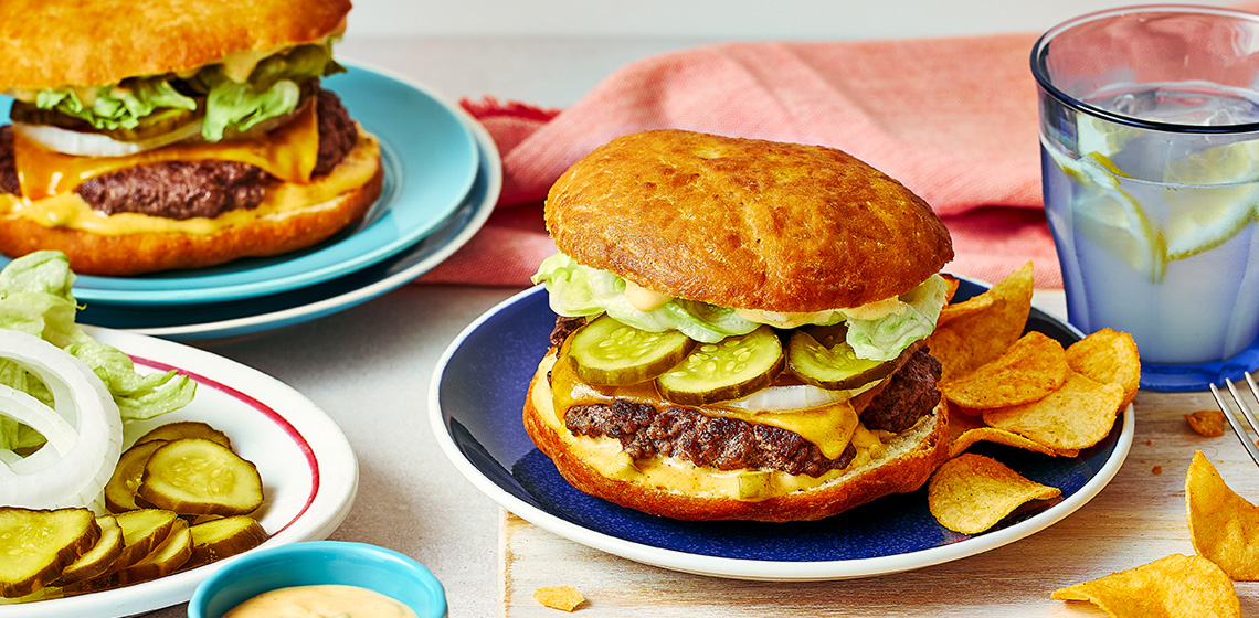Two bannock bison cheeseburgers with chips and sliced pickles and plates and a glass of lemon water.