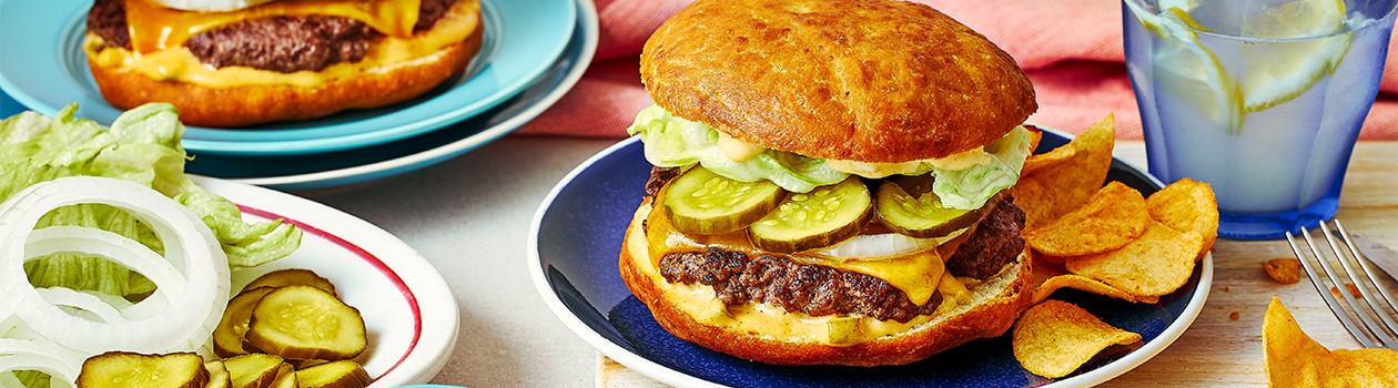 A bannock bison cheeseburger with chips and sliced pickles and plates and a glass of lemon water.