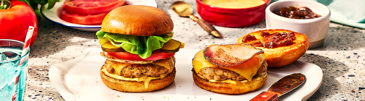 Pork and peameal bacon burger stacked high in a brioche bun, topped with pickles, tomato and cheddar cheese.