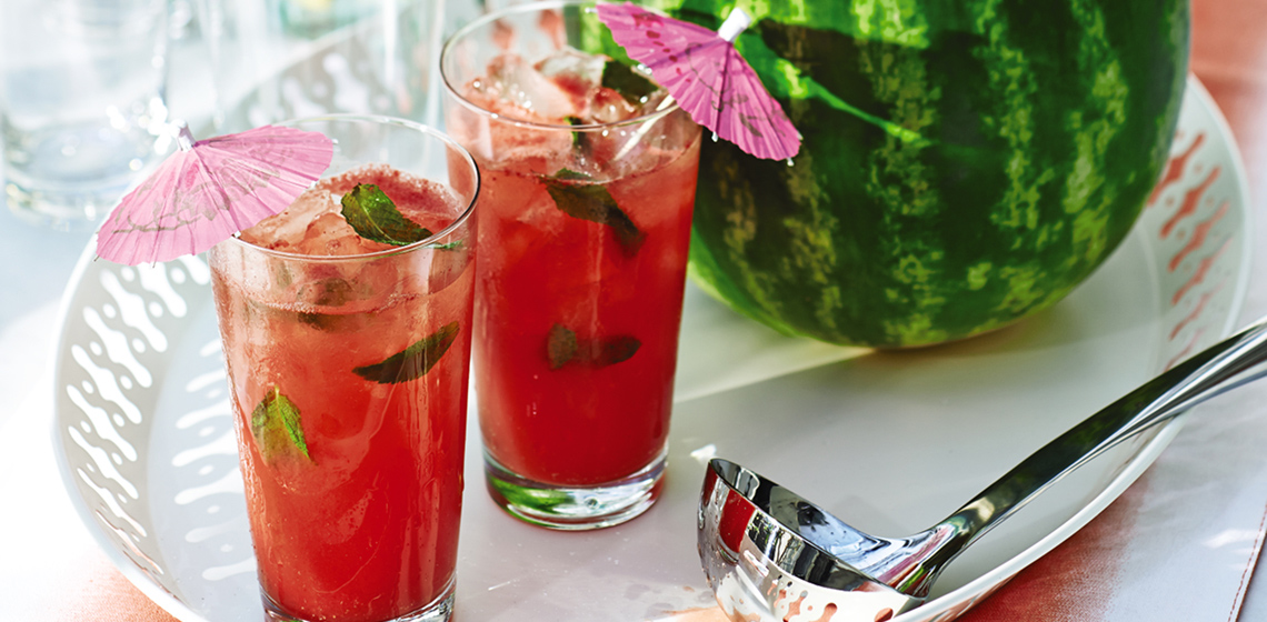 A white serving platter with watermelon rum punch in a hollowed-out watermelon and two tall glasses, and a silver serving ladle.