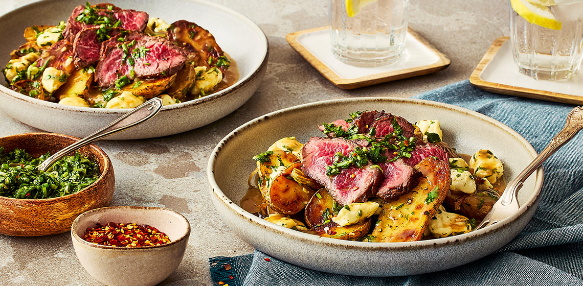 Two bowls of Argentinian poutine with two glasses of lemon water, a bowl of the chimichurri, and another small bowl with red pepper flakes.