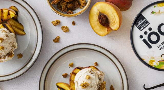 Two plates of Grilled Peach Sundaes made with iÖGO vanilla frozen yogurt, garnished with granola and served alongside fresh peaches.