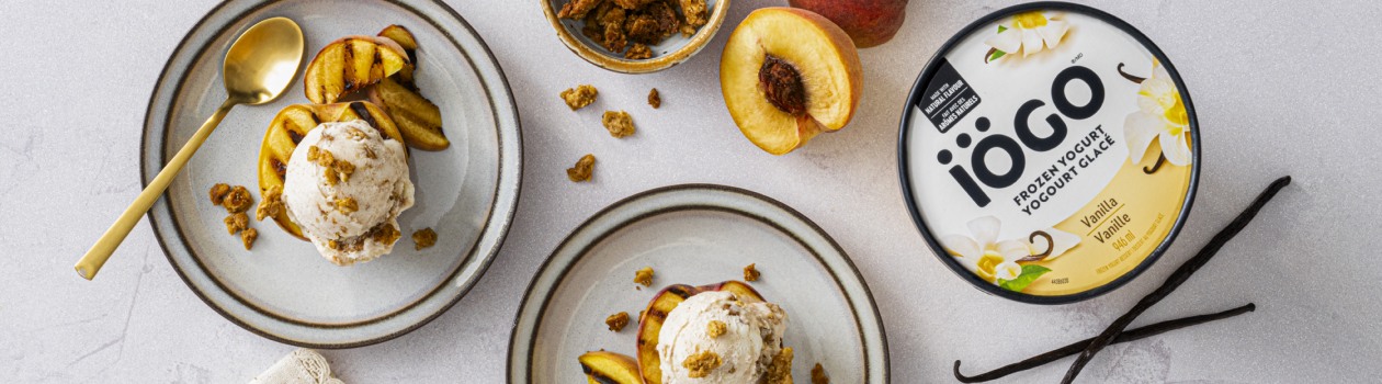 Two plates of Grilled Peach Sundaes made with iÖGO vanilla frozen yogurt, garnished with granola and served alongside fresh peaches.