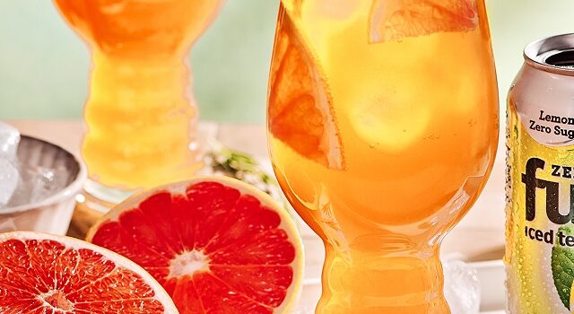 Lemon iced tea is served in a tall glass with ice and rosemary with two half sliced grapefruit on the left side.