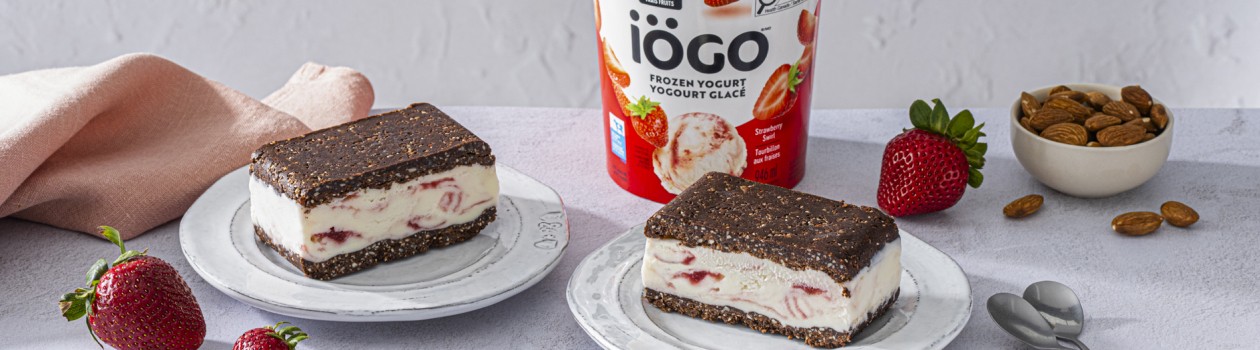 Two plates with No-Bake Brownie Frozen Yogurt Sandwiches made with iÖGO Strawberry Swirl Frozen Yogurt. The dessert is surrounded by fresh strawberries, almonds in a small bowl, and a tub of frozen yogurt in the center.