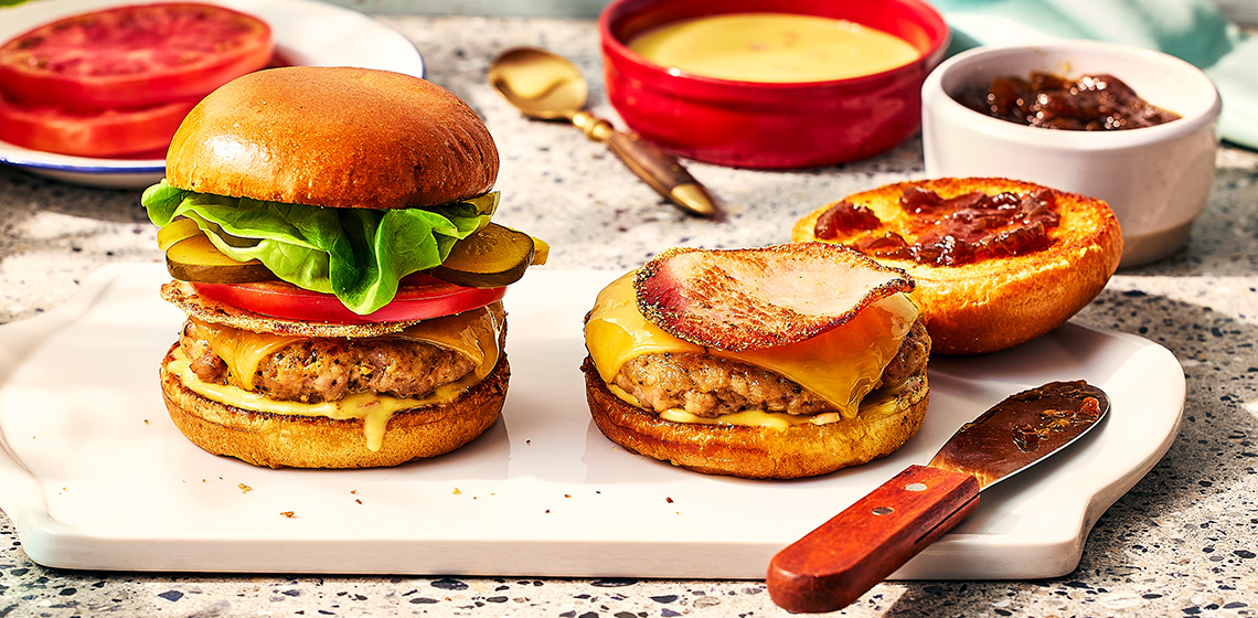 Pork and peameal bacon burger stacked high in a brioche bun, topped with pickles, tomato and cheddar cheese. 
